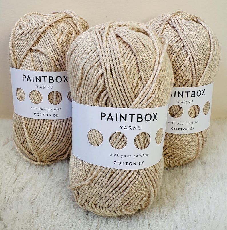 May include: Three beige yarn skeins with white labels that read "PAINTBOX YARNS" and "COTTON DK". The yarn is tightly wound, and the labels feature a series of circles. The yarn is displayed on a soft, white surface.
