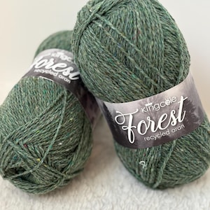 Op de afbeelding: Twee bollen groen garen met kleurrijke spikkels. Het garen is gelabeld "King Cole Forest recycled aran". Het garen heeft een gedempte groene kleur, met een licht getextureerde uitstraling. Het garen is strak opgerold tot een bol.