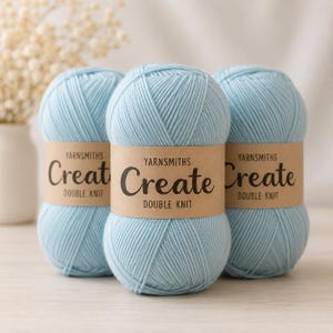 Puede incluir: Tres ovillos de lana azul claro con etiquetas de papel marrón que dicen "YARNSMITHS Create DOUBLE KNIT". La lana está dispuesta en un primer plano, con un fondo borroso de flores blancas en un jarrón.