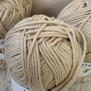 May include: Close-up of several beige yarn balls. Each ball is tightly wound and has a textured appearance. White labels with the text "PAINTBOX YARNS" are wrapped around the yarn balls. The yarn appears to be a natural fiber, suitable for knitting or crochet projects.