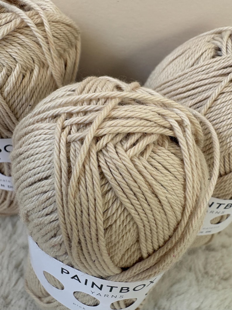 May include: Close-up of several beige yarn balls. Each ball is tightly wound and has a textured appearance. White labels with the text "PAINTBOX YARNS" are wrapped around the yarn balls. The yarn appears to be a natural fiber, suitable for knitting or crochet projects.