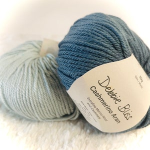 Op de afbeelding: Twee bollen garen, één blauw en één lichtblauw, met een label met de tekst "Debbie Bliss Cashmerino Aran Extra fine Merino Wool and Cashmere 50g 90m / 98yds"