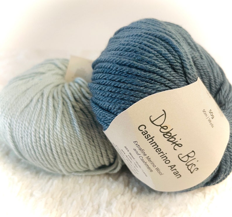 May include: Two skeins of yarn, one blue and one light blue, with a label that reads "Debbie Bliss Cashmerino Aran Extra fine Merino Wool and Cashmere 50g 90m / 98yds"