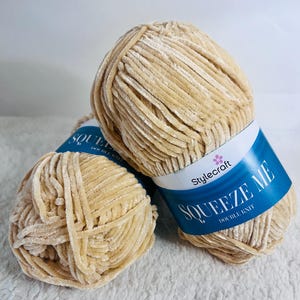 Puede incluir: Dos ovillos de hilo beige etiquetados "Squeeze Me Double Knit" de Stylecraft.