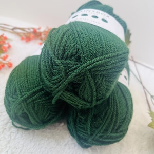 May include: Three balls of dark green yarn are stacked. The yarn is tightly wound, and the texture is visible. A white label with the text "PAINTBOX SIMPLY ARAN" is visible on one of the balls. The yarn is suitable for knitting.