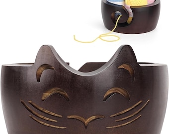 May include: A brown wooden yarn bowl shaped like a cat's face with a carved smile and whiskers. The bowl has a hole in the back for yarn to feed through. There are three balls of yarn in the bowl, one blue, one pink, and one yellow.