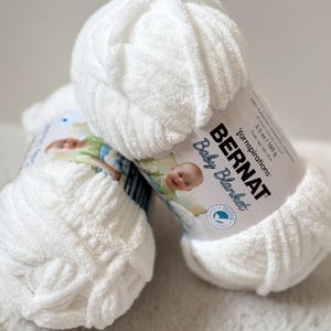 Pode incluir: Dois novelos de lã branca Bernat Baby Blanket. Cada novelo é rotulado com o nome da marca, nome do produto e peso. A lã tem uma textura macia e fofa, perfeita para cobertores de bebê. Cada novelo tem 100 g.
