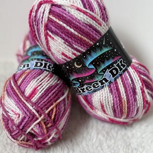 May include: Close-up of three skeins of yarn in shades of pink, white, and purple. Each skein has a black label with the text "Breeen DK" and a design of the aurora borealis. The yarn is arranged on a white, textured surface.