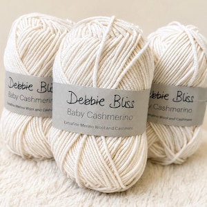 May include: Three ivory-colored yarn skeins are displayed, each wrapped with a gray label. The labels read "Debbie Bliss Baby Cashmerino, Extrafine Merino Wool and Cashmere." The yarn is tightly wound, showing the texture of the fibers.