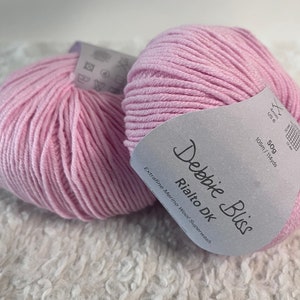 May include: Two balls of pink Debbie Bliss Rialto DK yarn, a superwash extrafine merino wool. The label reads "Debbie Bliss Rialto DK Extrafine Merino Wool Superwash 50g 105m/114yds Am US 6"