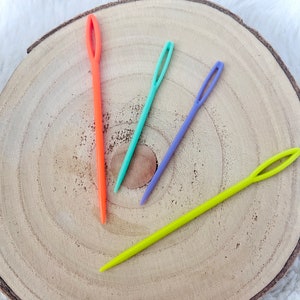 Puede incluir: Cuatro agujas de plástico de diferentes colores: naranja, azul claro, morado y amarillo. Las agujas están sobre una superficie de madera.