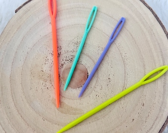 May include: Four plastic needles in different colors: orange, light blue, purple, and yellow. The needles are lying on a wooden surface.