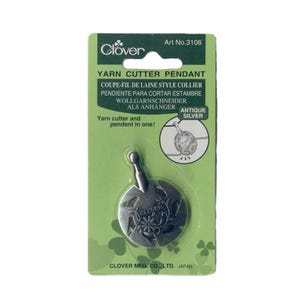 May include: Antique silver yarn cutter pendant by Clover. The pendant has a circular design with floral details and the Clover logo. The packaging is green with text in multiple languages, including "Yarn Cutter Pendant."