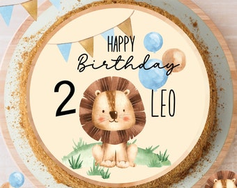 Könnte beinhalten: Ein runder Geburtstagskuchen mit einer Löwenillustration, der Zahl 2 und dem Namen "Leo". Der Kuchen ist mit blauen und goldenen Akzenten verziert, darunter Luftballons und ein Banner mit der Aufschrift "Happy Birthday". Der Kuchen steht auf einem Holzservierteller.