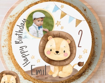 Könnte beinhalten: Ein runder weißer Kuchen mit einem Löwenmotiv, blauer und goldener Girlande, Sternen und dem Text "Happy Birthday Emil" und der Zahl "2". Ein Foto eines kleinen Jungen befindet sich in einem Kreis auf dem Kuchen.