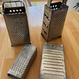 May include: Four vintage metal graters on a wooden table. Two are rectangular with handles, one has the word "BRONCO" on it. The other two are flat, rectangular graters. All have a worn, aged appearance.