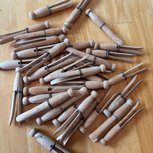 May include: A collection of light brown wooden clothespins, each featuring a two-pronged design held together by a thin, dark metal wire coil. The pins are scattered in a pile, showing their natural wood grain and smooth, rounded handle ends.