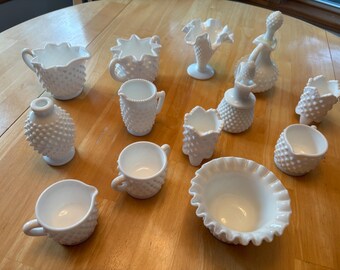 May include: A collection of twelve white milk glass pieces featuring hobnail textures, including various pitchers, vases, a decanter with a stopper, and a ruffled bowl. Each piece displays a distinct shape, ranging from small creamers to tall vessels.