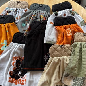 May include: A collection of hanging kitchen towels with various Halloween and fall-themed designs. The towels feature images of Mickey Mouse, pumpkins, and skeletons. Each towel has a quilted top with a button closure, and the colors include orange, black, white, and green.