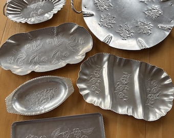 May include: A collection of vintage aluminum serving trays and dishes. The trays vary in shape, including oval, rectangular, and round with handles. Each piece features embossed floral designs and a silver finish, suitable for serving food.