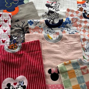 May include: A collection of assorted fabric bibs featuring various patterns including red and white stripes with Mickey and Minnie Mouse, pink with a Minnie Mouse silhouette, checkered prints, and Halloween-themed designs with pumpkins and ghosts.