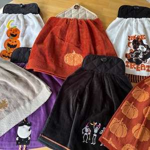 May include: A collection of seven hanging kitchen towels featuring various Halloween and autumn designs, including pumpkins, skeletons, a ghost, and a mouse character with the text Trick or Treat, all attached to quilted fabric tops with buttons.