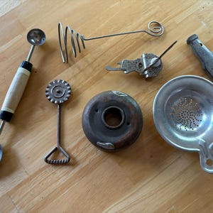 May include: A collection of vintage kitchen tools, including a melon baller, a cookie cutter, a whisk, a strainer, a springform pan, and a can opener.