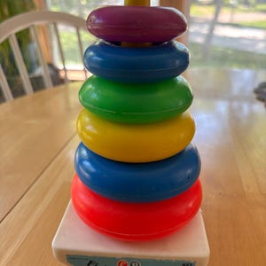 May include: A vintage Fisher-Price Toys stacking ring toy. The toy features a white base with a red, blue, yellow, green, and purple ring stack. The top is a yellow peg. The base has the Fisher-Price Toys logo and the text "East Aurora, N.Y."