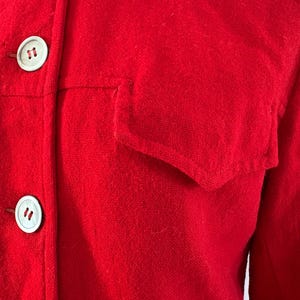 May include: Close-up of a bright red jacket with two white buttons and a pocket. The jacket is made of a textured fabric, and the buttons are round with a subtle design. The overall style is classic and simple.