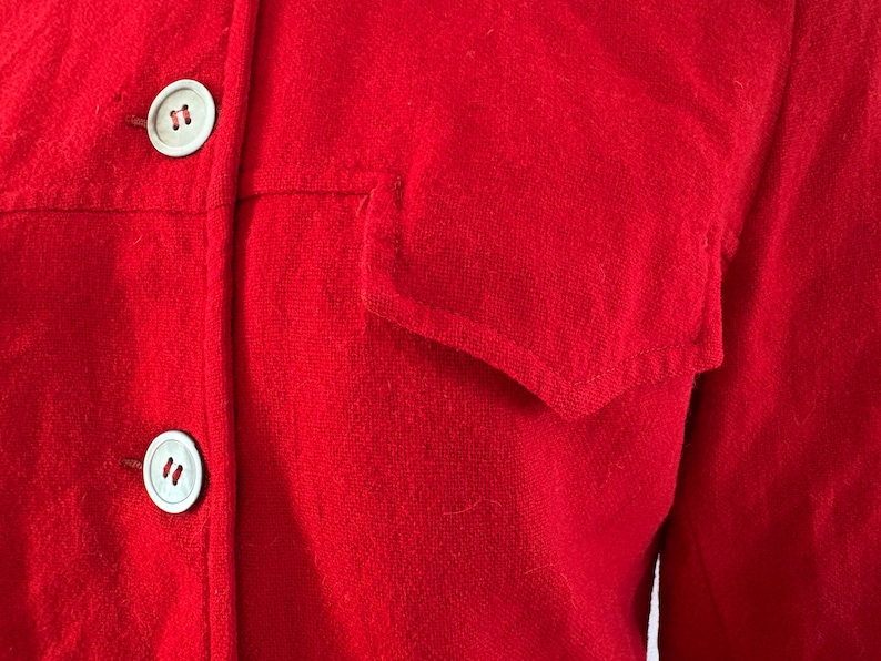 May include: Close-up of a bright red jacket with two white buttons and a pocket. The jacket is made of a textured fabric, and the buttons are round with a subtle design. The overall style is classic and simple.
