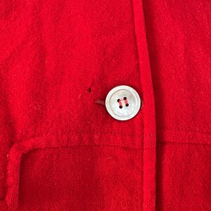 May include: Close-up of a bright red jacket with a large, round, pearlescent button. The jacket has a simple design with a single button closure and a small pocket detail. The fabric appears to be a soft, textured material.