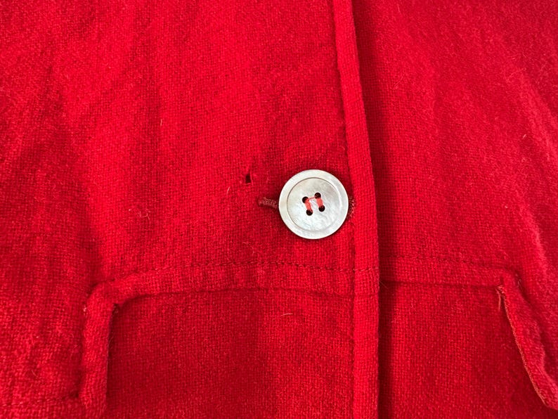 May include: Close-up of a bright red jacket with a large, round, pearlescent button. The jacket has a simple design with a single button closure and a small pocket detail. The fabric appears to be a soft, textured material.