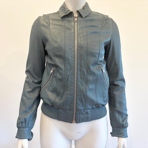 May include: A teal-colored leather jacket with a zip-up front, two zippered pockets, and a ribbed hem and cuffs. The jacket has a collared neckline and vertical stitched details on the front and sleeves.