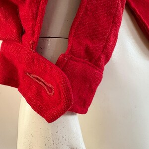 May include: Close-up of a bright red garment, possibly a jacket or coat, with a textured weave. The fabric is a solid, vibrant red. The image focuses on the sleeve and a buttoned cuff, showcasing the tailored details and construction of the clothing item.