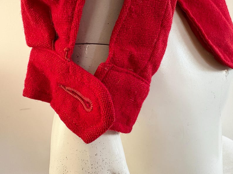 May include: Close-up of a bright red garment, possibly a jacket or coat, with a textured weave. The fabric is a solid, vibrant red. The image focuses on the sleeve and a buttoned cuff, showcasing the tailored details and construction of the clothing item.