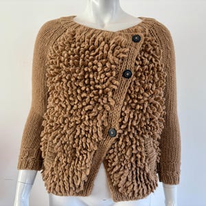 May include: A tan-colored, textured knit cardigan with a unique, raised design on the front. The sweater features a round neckline, long sleeves, and three dark buttons. The overall style is a cozy, textured knitwear.