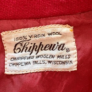 May include: Close-up of a red fabric with a sewn-in label. The label reads "100% Virgin Wool" and features the brand name "Chippewa" in a script font. Below, it states "Chippewa Woolen Mills, Chippewa Falls, Wisconsin."