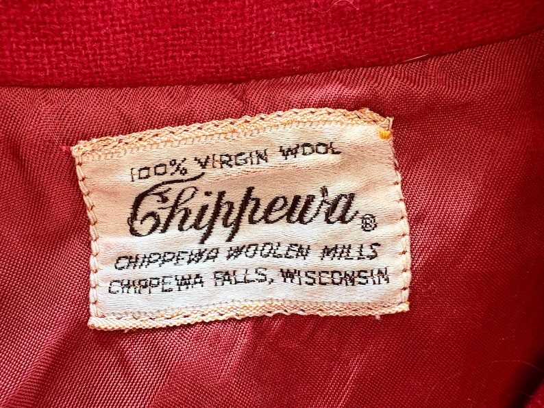 May include: Close-up of a red fabric with a sewn-in label. The label reads "100% Virgin Wool" and features the brand name "Chippewa" in a script font. Below, it states "Chippewa Woolen Mills, Chippewa Falls, Wisconsin."