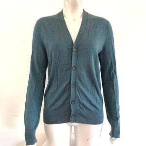 May include: Teal-colored cardigan sweater with a V-neck and button-up front. The long-sleeved sweater is displayed on a mannequin against a white background. The sweater has a ribbed hem and cuffs.