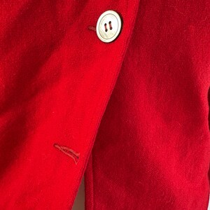 May include: Close-up of a vibrant red blazer. The blazer features a large, round, off-white button with two small holes. The fabric has a soft texture. A buttonhole is visible on the lapel. The blazer is a solid color.