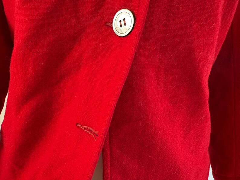 May include: Close-up of a vibrant red blazer. The blazer features a large, round, off-white button with two small holes. The fabric has a soft texture. A buttonhole is visible on the lapel. The blazer is a solid color.