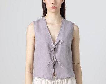 May include: A sleeveless, lavender-colored vest with a V-neck and tie closures. The vest is paired with off-white linen pants. The outfit is simple and elegant, suitable for warm weather. The vest is made of a lightweight fabric.