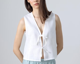 May include: A white sleeveless top with a deep V-neck and tie closures. The top is paired with light blue pants. The outfit is made of a lightweight fabric, perfect for warm weather. The model is wearing the outfit.