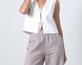 May include: A sleeveless white linen top with a V-neck and tie closure, paired with wide-leg, light purple linen pants. The outfit is simple and elegant, suitable for warm weather. The fabric appears lightweight and breathable.