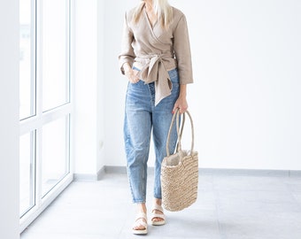 May include: A woman wearing a beige wrap top, light blue jeans, and sandals, holding a woven straw tote bag. The outfit is casual and stylish. The bag is approximately 14 inches tall.