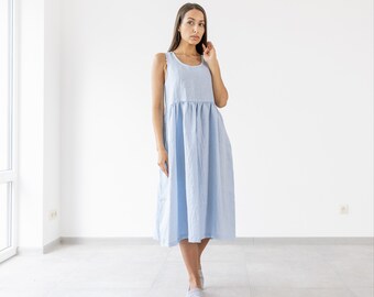 May include: A woman wearing a light blue linen dress with a gathered waist. The dress is sleeveless and falls to her knees. She is standing in a white room with a window in the background.