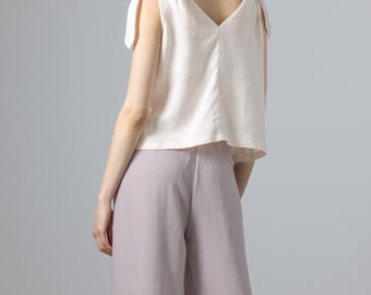 Puede incluir: Una blusa de lino beige claro con tirantes anudados y cuello en V. La blusa se combina con pantalones anchos de color lila claro. El conjunto es sencillo y elegante, adecuado para ocasiones informales o semiformales.