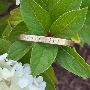 May include: A gold-colored metal cuff bracelet with the words "choose joy" stamped across the front. The bracelet rests on a bed of green leaves and white flowers, creating a natural and serene setting. The bracelet is a simple, yet elegant piece of jewelry.