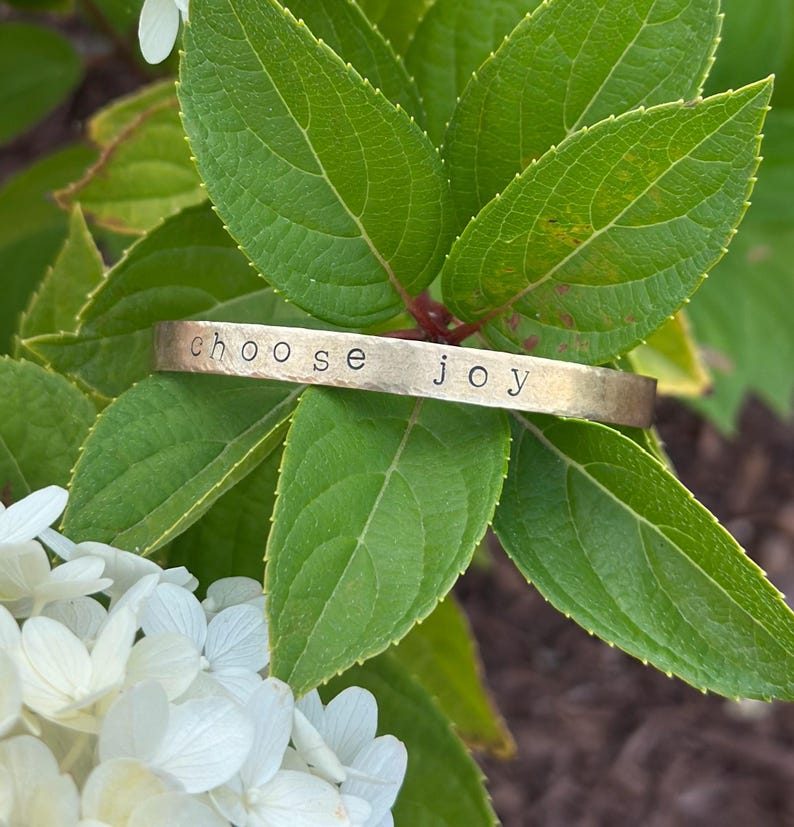 May include: A gold-colored metal cuff bracelet with the words "choose joy" stamped across the front. The bracelet rests on a bed of green leaves and white flowers, creating a natural and serene setting. The bracelet is a simple, yet elegant piece of jewelry.