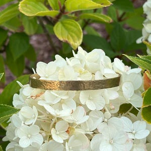 May include: A hammered gold-tone bracelet rests on a bed of white hydrangea flowers. The bracelet has a textured surface and a simple, elegant design. Green leaves and flowers are in the background.